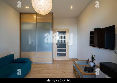 Contemporary interior of living room in modern apartment. Laptop and printer. Sliding door wardrobe. TV on wall. Wooden door. Stock Photo