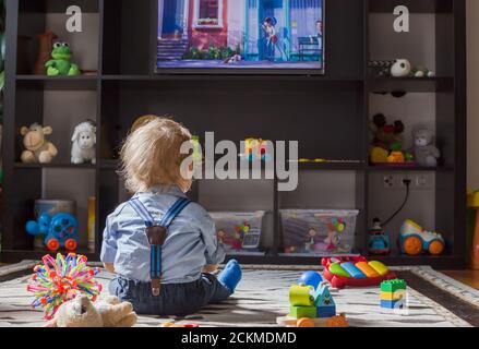 Cute baby boy and his teddy bear watching TV sitting on the floor covered with toys, at home Stock Photo