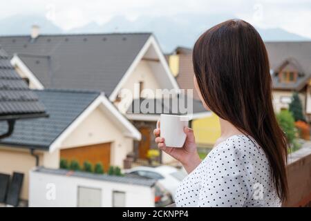 Woman standing with a white cup on the balcony and dreamily watching on the cloudy mountain hill. Morning in the countryside. Modern houses near the Stock Photo