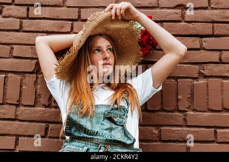 Portrait of young, teen ginger girl in blue jeans with suspenders and a hat. Hands above her head holding bouquet of red flowers Stock Photo