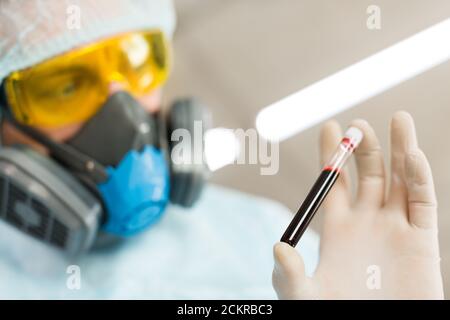 Researcher in protective wear is carefully looking at a blood sample in a test tube. Creating a vaccine. Close up photo. Focus on test tube. Stock Photo