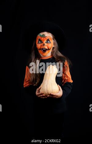a girl in a black hat and with pumpkin makeup on her face holds a pumpkin in her hands and celebrates Halloween  Stock Photo