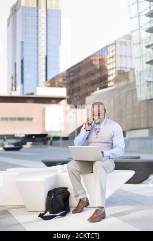 Black businessman talking to his smartphone while driving his laptop on the street. Concept of new ways of business. Space for text. Stock Photo