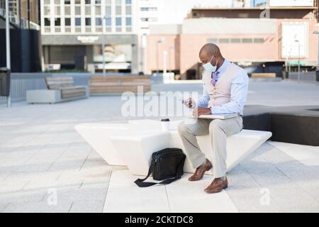 Black businessman sitting on the street with his work tools. He is wearing a medical mask as a precaution against the Covid-19 pandemic. Stock Photo