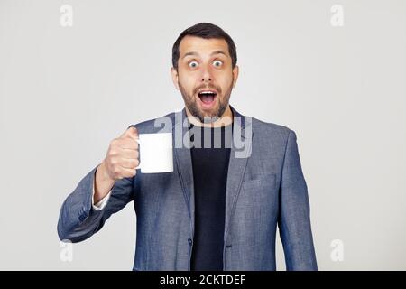 Young businessman man with a beard in a jacket, holding a cup of coffee, scared in shock with a surprised face, scared and excited with an expression of fear. Portrait of a man on a gray background. Stock Photo