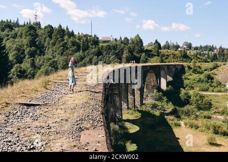 Young tourist girl walking on ancient bridge. Woman sitting on viaduct with old railway tracks near green hill of mountain forest. Locat travel Stock Photo