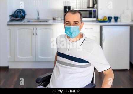 Smiling Young Handicapped Man Sitting On Wheelchair In Kitchen.  Young man wearing face mask sitting infront of kitchen. Focus on his face. Stock Photo