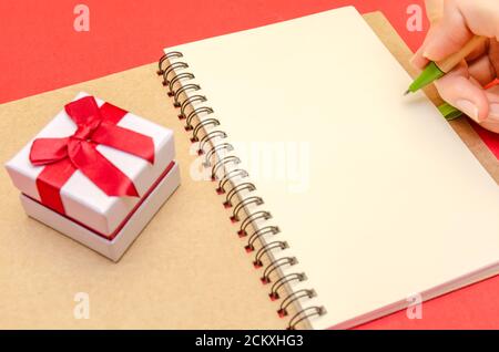 hand of a girl who is going to draw on a blank sheet in a notebook, next to a gift box on a red background. The concept of Valentine's Day, holiday Stock Photo