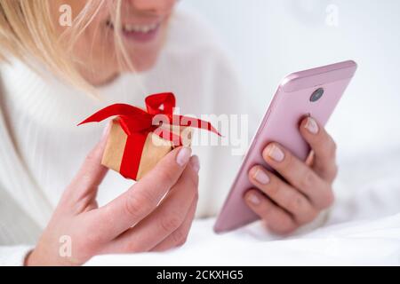 Christmas online shopping. Female buyer makes order on mobile phone. Woman buy presents, prepare to xmas, gift box in hand. Winter holidays sales  Stock Photo
