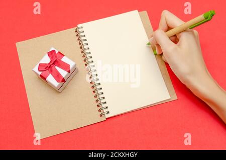 hand of a girl who is going to draw on a blank sheet in a notebook, next to a gift box on a red background. The concept of Valentine's Day, holiday Stock Photo