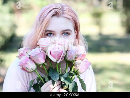 Beautiful girl with blue eyes sniffs pink roses, outdoors. Summertime. Stock Photo