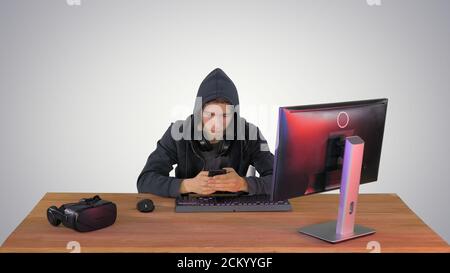 Hooded hacker using laptop and smartphone on gradient background Stock Photo