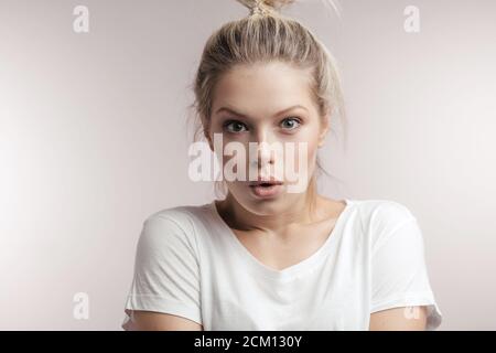 Studio portrait of attractive surprised astonished blonde woman with hair bun, looking at camera in full disbelief, having shocked expression on her f Stock Photo