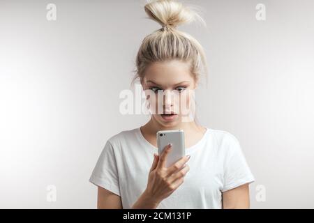 Shocked young caucasian woman looks with surprised amazed expression at her smart phone and bated breath, reads reminder and realizes that she forgot Stock Photo