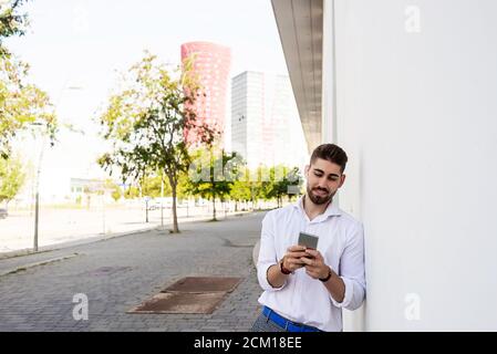 Young bearded man leaning against a white wall using the mobile phone Stock Photo
