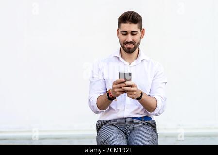Young bearded man sitting against a white wall using the mobile phone Stock Photo