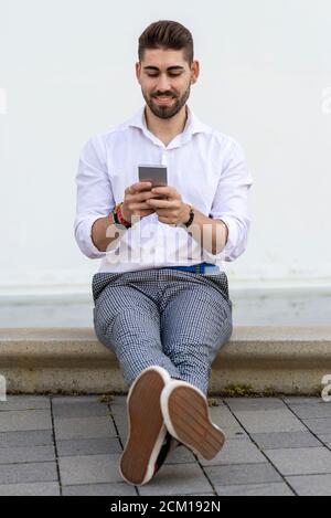 Young bearded man sitting against a white wall using the mobile phone Stock Photo
