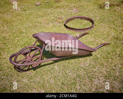 An old timey rusted metal wheel barrow sits in the grass in the sun. Stock Photo