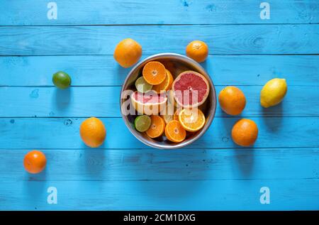 Slices of chopped fresh juicy citrus fruits as oranges, mandarines, lemons in a bowl on a blue wooden background. Several whole citrus fruits lying ne Stock Photo