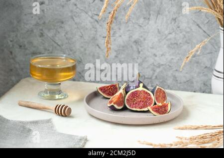 fresh ripe slices of figs on stylish beige ceramic plate, bowl with honey and honey stick, gray napkin and bouquet of dried flowers. cozy healthy Brea Stock Photo