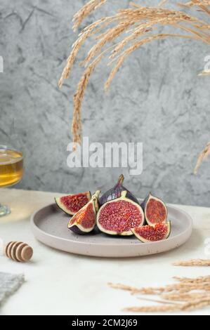 fresh ripe slices of figs on stylish beige ceramic plate, bowl with honey and honey stick, gray napkin and bouquet of dried flowers. cozy healthy Brea Stock Photo