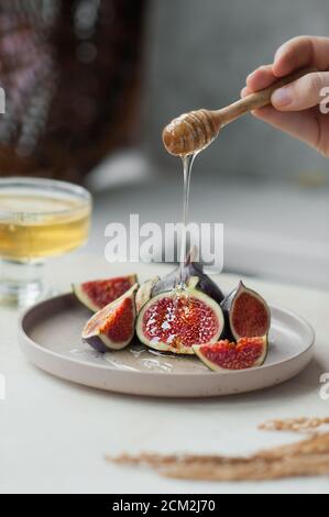 honey drips from honey wooden stick to pieces of fresh ripe figs on stylish ceramic plate, bowl of honey and dried flowers on the background, selectiv Stock Photo