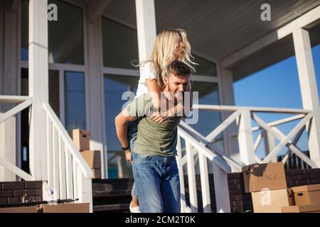 Husband giving wife piggyback near new home outside. cardboard boxes near steps Stock Photo