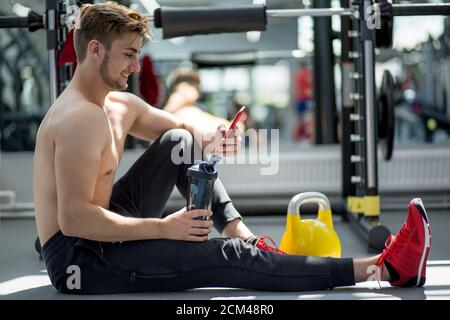 Athlete wearing red sneakers sitting on the floor in gym with bottle of water and using smartphone Stock Photo