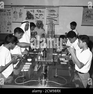 1960s, historical, male pupils in a science laboratory at a secondary school in Jeddah, Saudi Arabia, with an arab male teacher in traditional thawb or robe and headdress instructing them at the work bench. The boys are conducting experiments and are using bunsen or gas burners, standard equipment in schools science lessons in this and previous eras and used for heating, burning and sterilising materials and liquids. Stock Photo