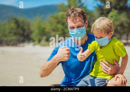 Father squatting holding son playing fists with thumbs up, wearing face blue mask protect coronavirus walking nature sand sea beach near forest park Stock Photo