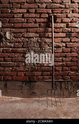Old pitchfork leaning against red brick wall, old and weathered on a farm, indoor light coming from outside Stock Photo