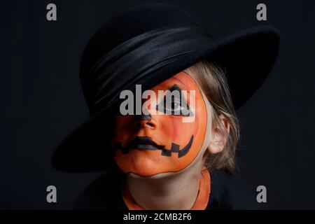 sad child with a pumpkin pattern on his face in the black hat on a black background, Halloween and looks like a Joker Stock Photo