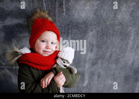 Close-up portrait of a cute little kid girl in a red hat and scarf hugging a knitted toy teddy bear, children safety and protection concept, child loo Stock Photo