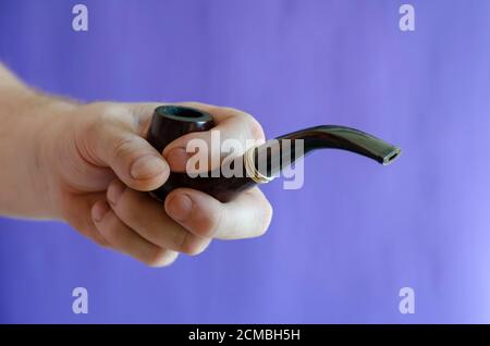 Smoking pipe in hand against the background of a blue wall. An adult male's right hand holds a smoking pipe made from heather or briar root. Monochrom Stock Photo
