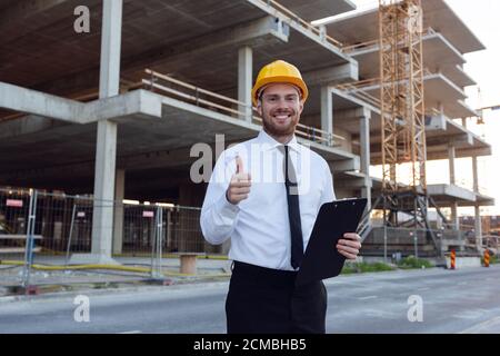 Businessman wearing Hardhat Holding Clipboard in hands Showing Thumb Up. Builder Controlling House Building. Man Smilling near Construction Works Stock Photo