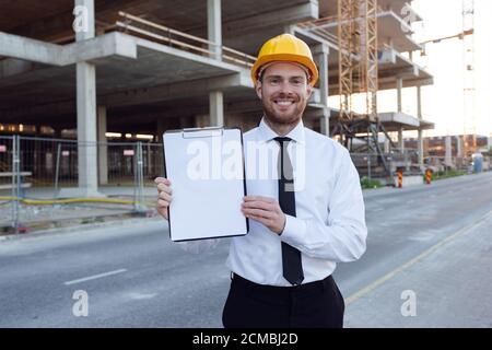 Businessman wearing Hardhat Showing Clipboard. Builder Controlling House Building. Man Smilling near Construction Works Stock Photo