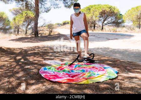 girl with a mask on her face and sunglasses inflating a colorful Inflatable Mattress Lollipop with her foot and a bomb to enjoy the vacations in the m Stock Photo
