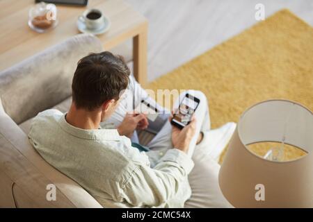 Rear view of young man sitting on sofa with mobile phone and shopping online using his credit card Stock Photo