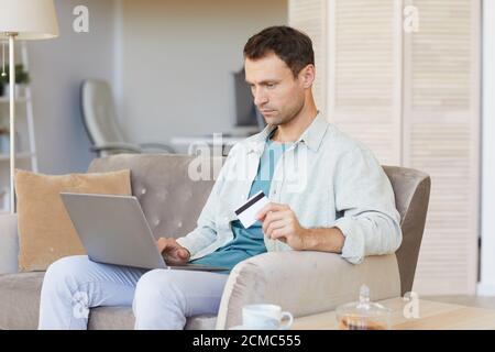 Young serious man sitting on sofa with laptop and paying bills online with credit card at home Stock Photo
