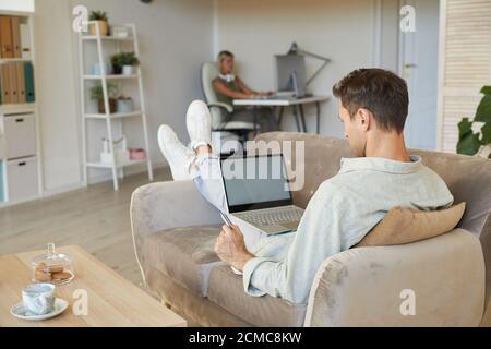 Rear view of young man lying on sofa and working online on laptop in the room at home Stock Photo