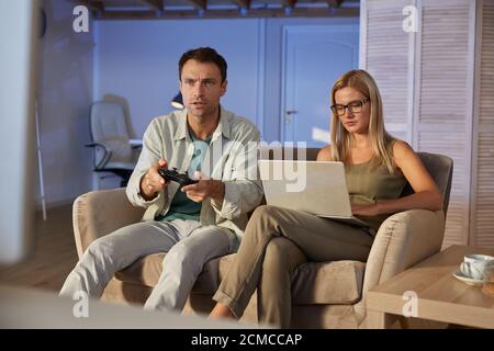 Young woman sitting on sofa and working on laptop while her boyfriend playing in computer games they sitting at home Stock Photo
