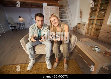 Young man playing together with his girlfriend in computer game they sitting on sofa and using joysticks in the living room Stock Photo