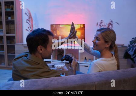 Young happy couple sitting on sofa and giving high-five to each other while playing the video game at home Stock Photo
