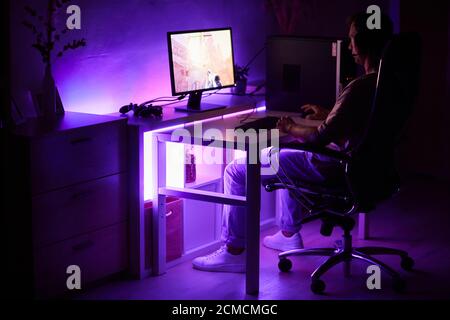 Gamer sitting at the table in front of computer monitor and playing computer game in dark room at home Stock Photo