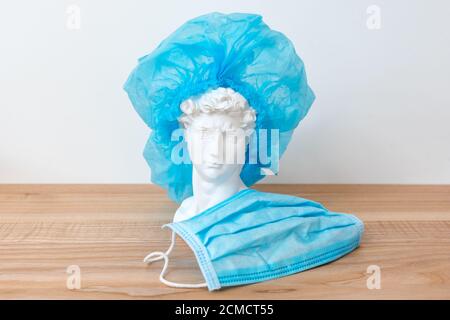 Gypsum copy of David's head in surgical cap. David plaster bust, ancient sculpture, statue on wooden table with medical mask. Medicine concept Stock Photo