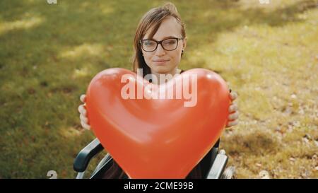 Beautiful young woman in the wheelchair holding heart shaped balloon in the park. High quality photo Stock Photo