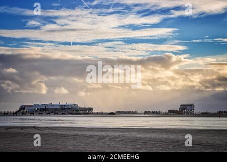 Panoramic view of stilt houses and wooden seabridge at low tide north sea beach during sunset in Sankt Peter-Ording, Germany against dramatic sky Stock Photo