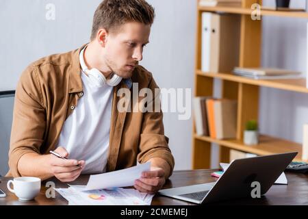 focused businessman with wireless headphones on neck holding document and looking at laptop Stock Photo