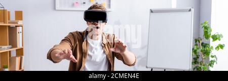 horizontal image of young businessman with open mouth using vr headset and gesturing at workplace Stock Photo