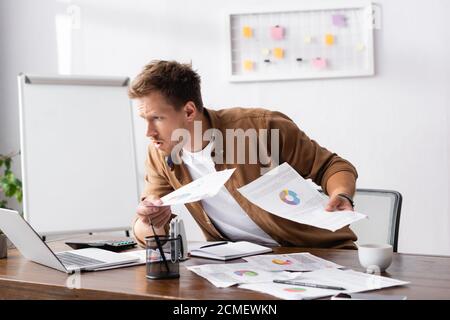 Selective focus of concentrated businessman holding papers and looking at laptop while working in office Stock Photo